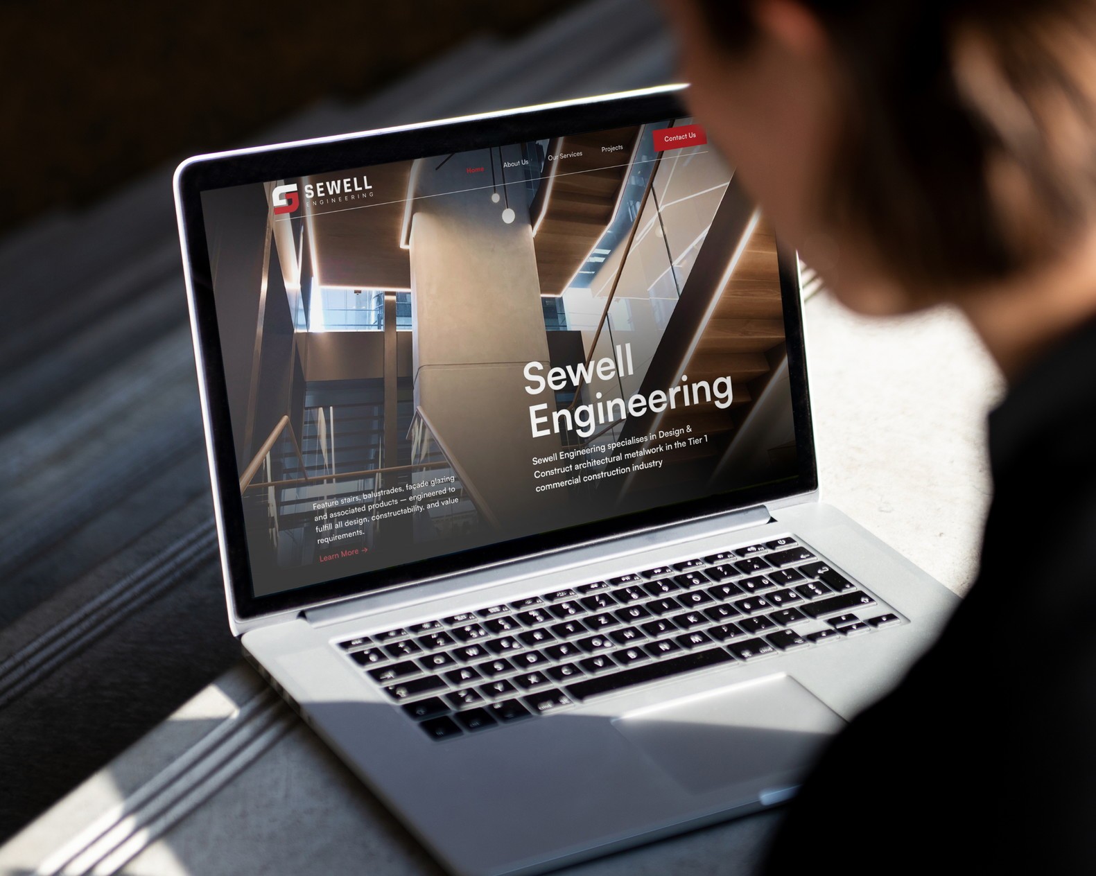 A photo is BrandVillage web design and development project of Sewell Engineering opened in a silver laptop on a wooden desk with a striped pattern. The laptop is open and the screen is visible.